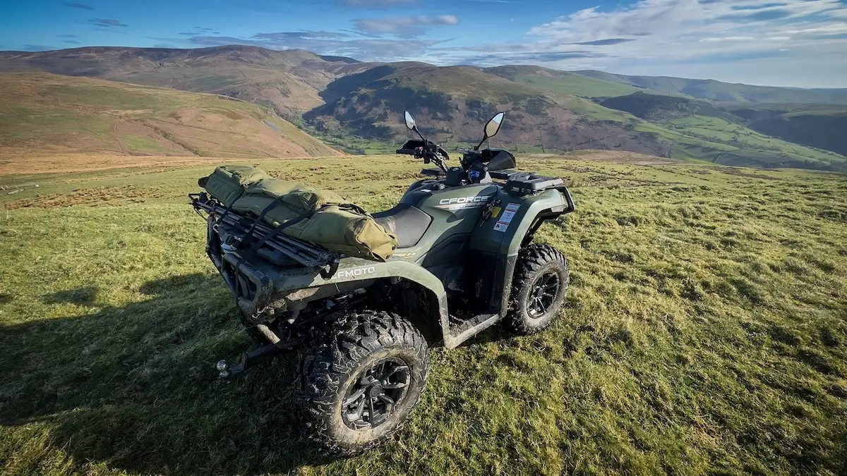 A cforce 520 four-wheel ATV with gear bags is parked on grassy terrain, overlooking rolling hills and valleys under a partly cloudy sky.