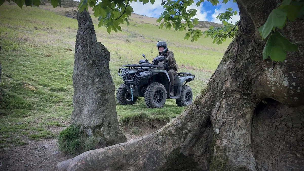 Person wearing a helmet rides a CForce 520 ATV on grassy terrain, framed by large rocks and tree trunks with green leaves in the foreground.
