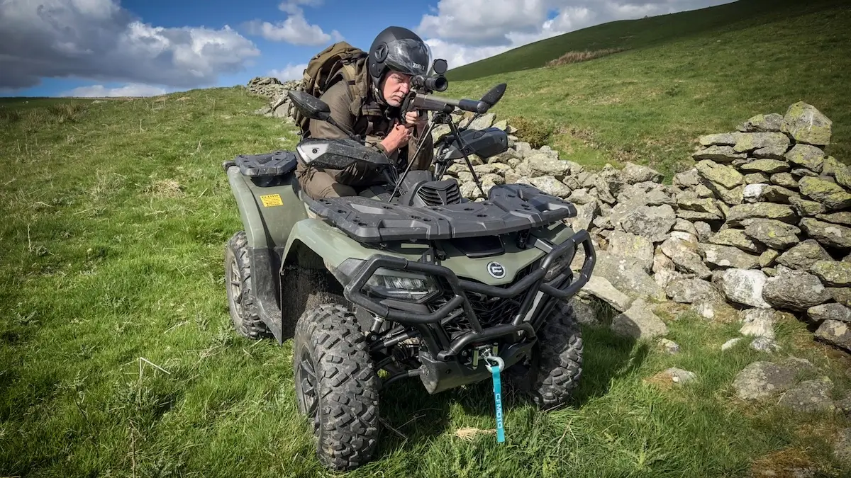 A person in tactical gear aims a rifle from the seat of a cforce 520 all-terrain vehicle parked on grassy terrain beside a stone wall.