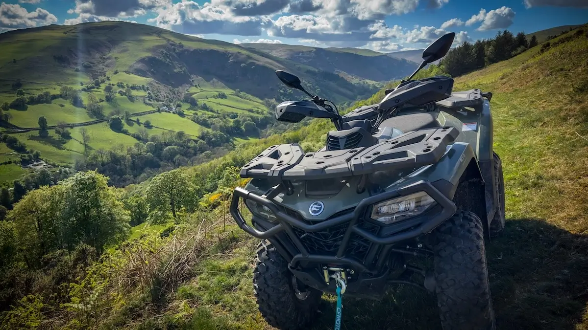 A black CForce 520 ATV is parked on a grassy hillside, overlooking a scenic valley with rolling green hills and scattered trees under a partly cloudy sky.