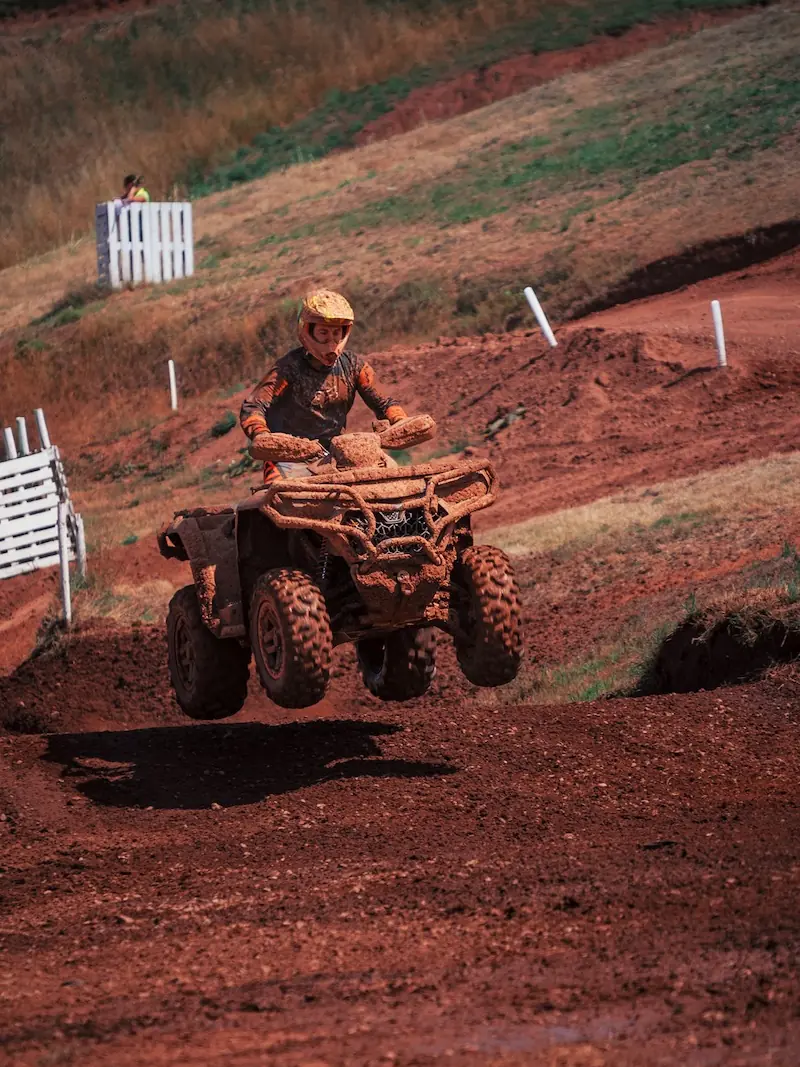 A rider on a CFMOTO CFORCE 1000 powers through the dirt track, launching airborne as mud flies—classic Mudbillys action in the 4x4 Class, with white fences and grassy areas framing the scene.