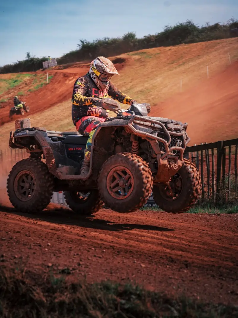 A person riding a CFMOTO CFORCE 1000 ATV off-road catches air over a dirt track, dust flying as they race in the Mudbillys 4x4 class, with another rider visible in the background.