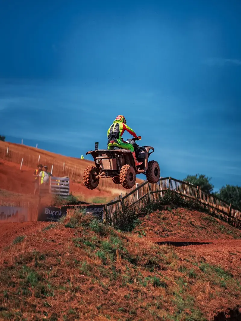A Mudbillys ATV rider wearing safety gear jumps over a dirt ramp on a motocross track under a clear blue sky, showcasing the power of the CFMOTO CFORCE 1000 in the 4x4 class.