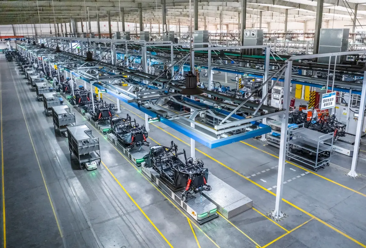A wide view of an automated factory floor shows rows of machinery and assembly lines, including equipment and vehicles like the CFMOTO CFORCE TECH GEN 4 Concept, all under bright industrial lighting.