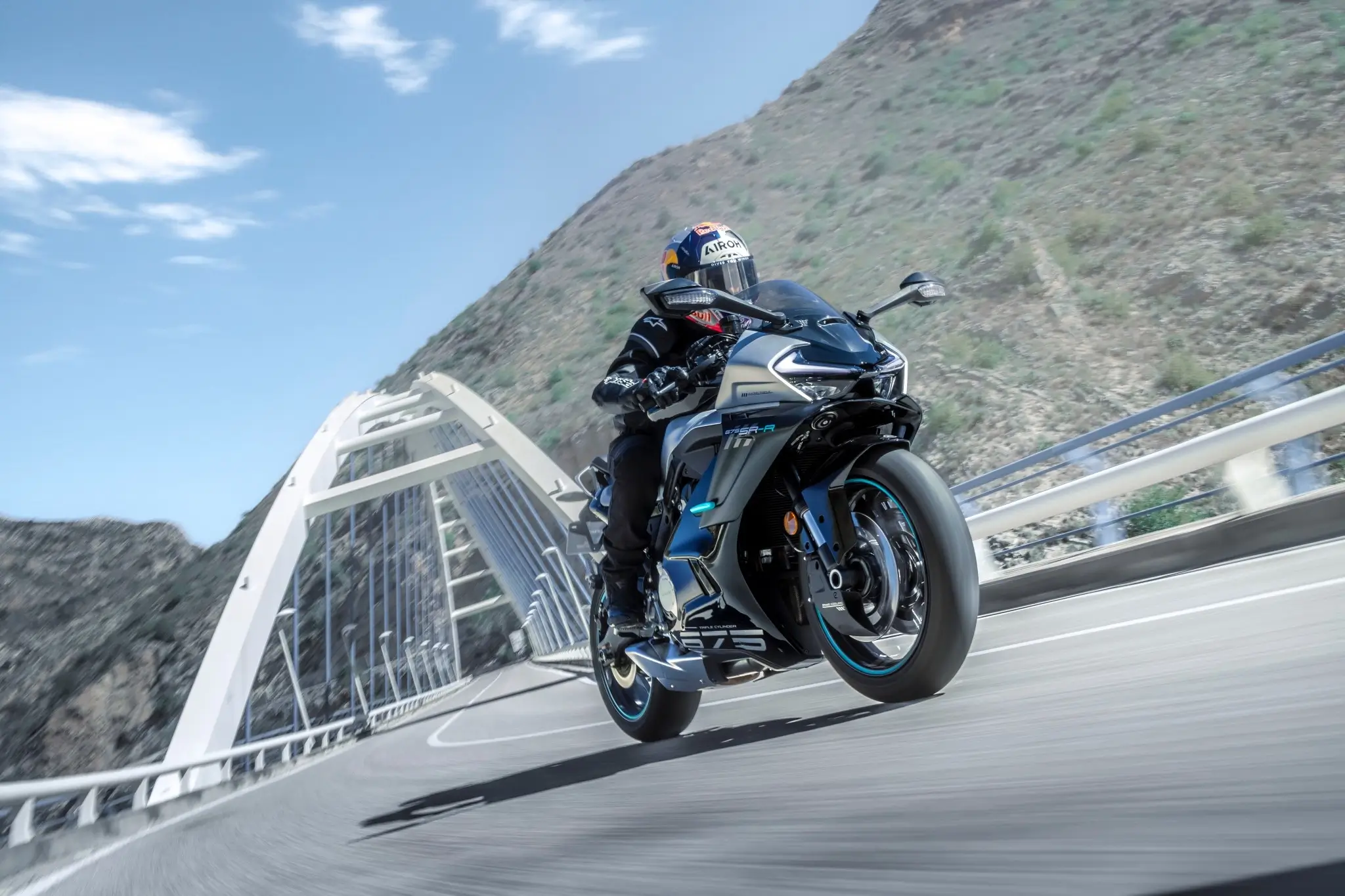 A person rides a 675SR-R sport motorcycle across a modern bridge on a winding road, with mountains in the background under a clear sky.