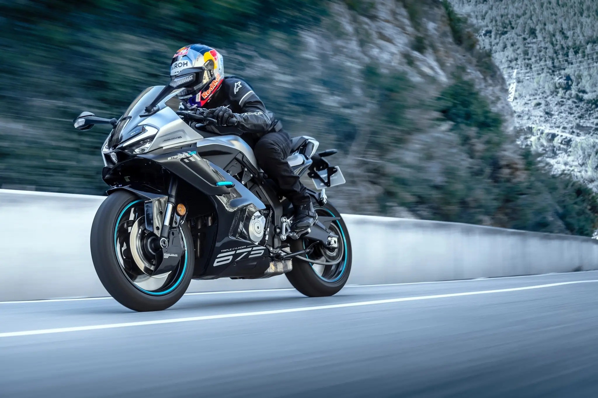 A motorcyclist in black gear rides a gray 675SR-R sport motorcycle at speed on a mountain road with blurred background.