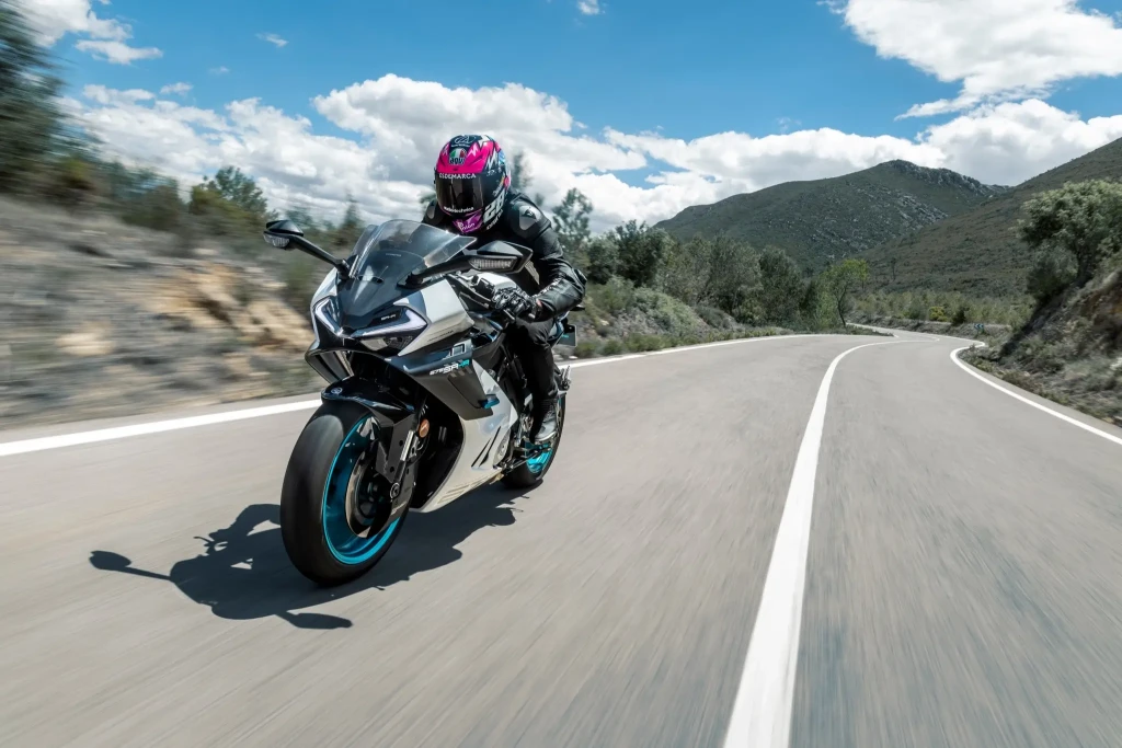 A person in black motorcycle gear and a pink-accented helmet rides a 675SR-R sport motorcycle on a winding road through a mountainous landscape under a partly cloudy sky.
