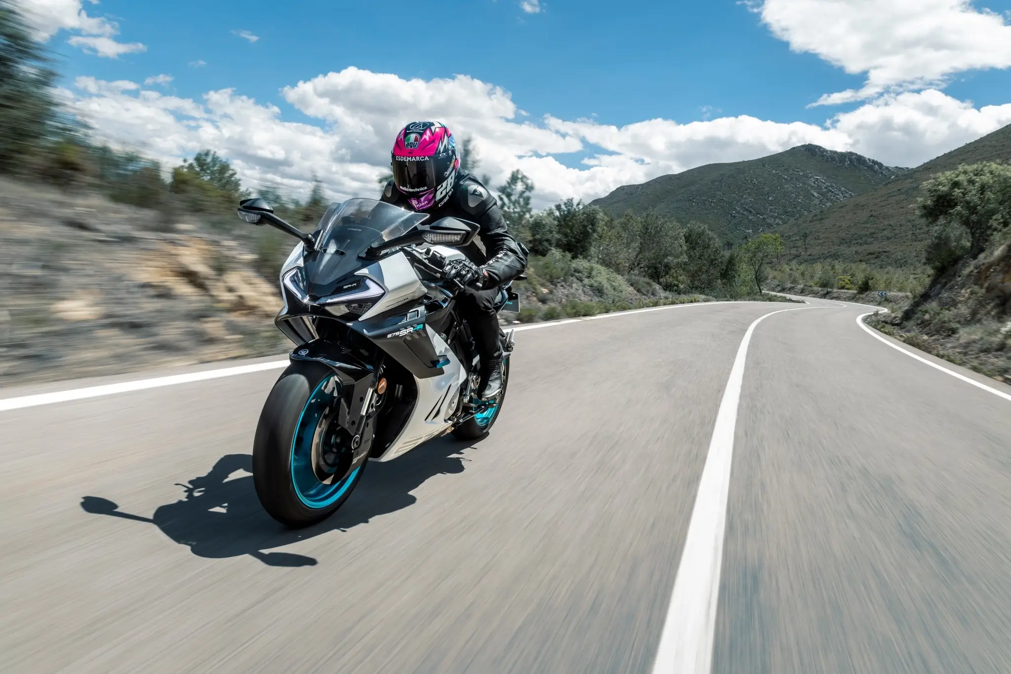A person in black motorcycle gear and a pink-accented helmet rides a 675SR-R sport motorcycle on a winding road through a mountainous landscape under a partly cloudy sky.