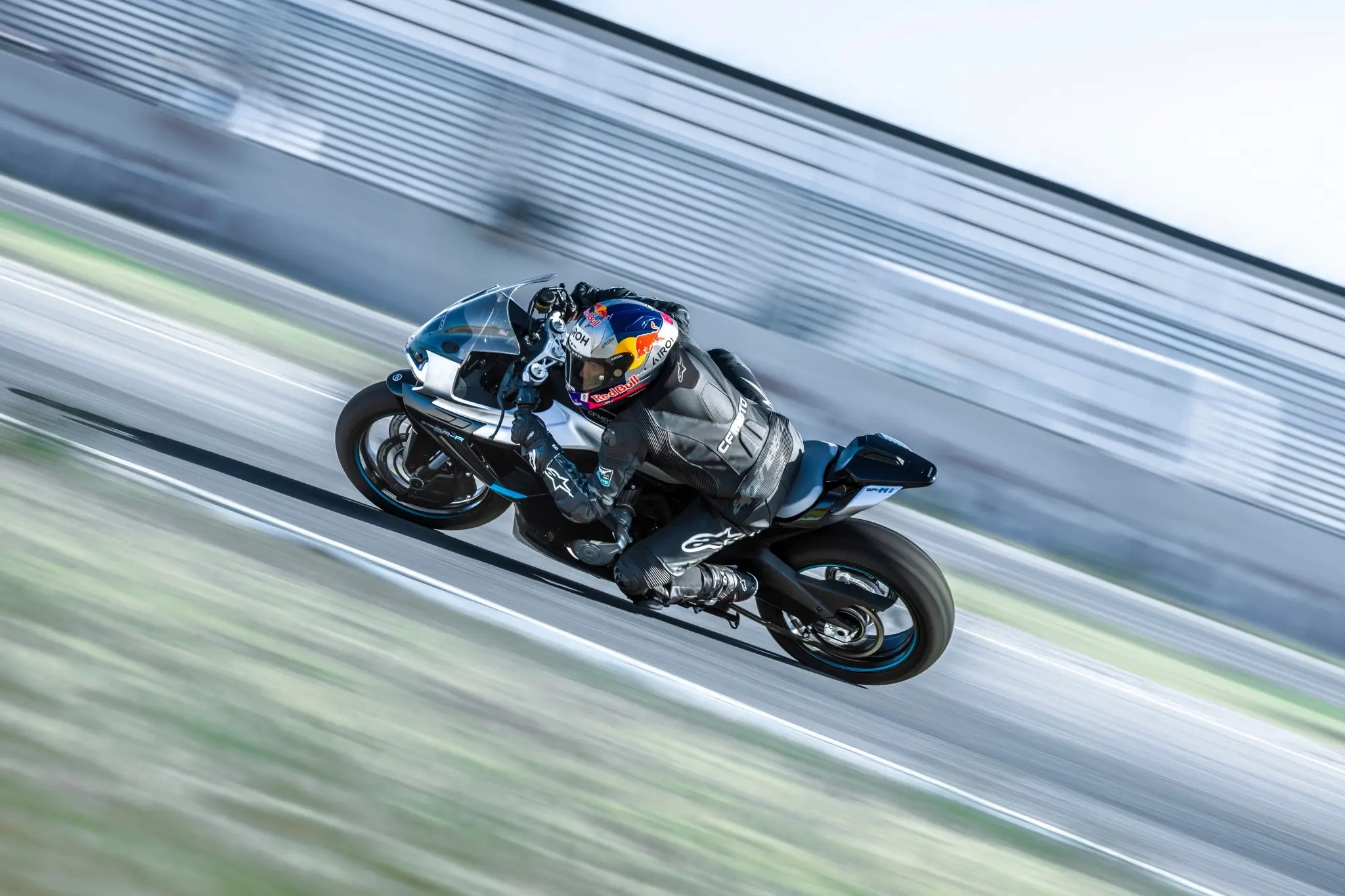 A motorcyclist in racing gear rides a blue 675SR-R sport bike at high speed around a track, leaning into a turn with a blurred background.