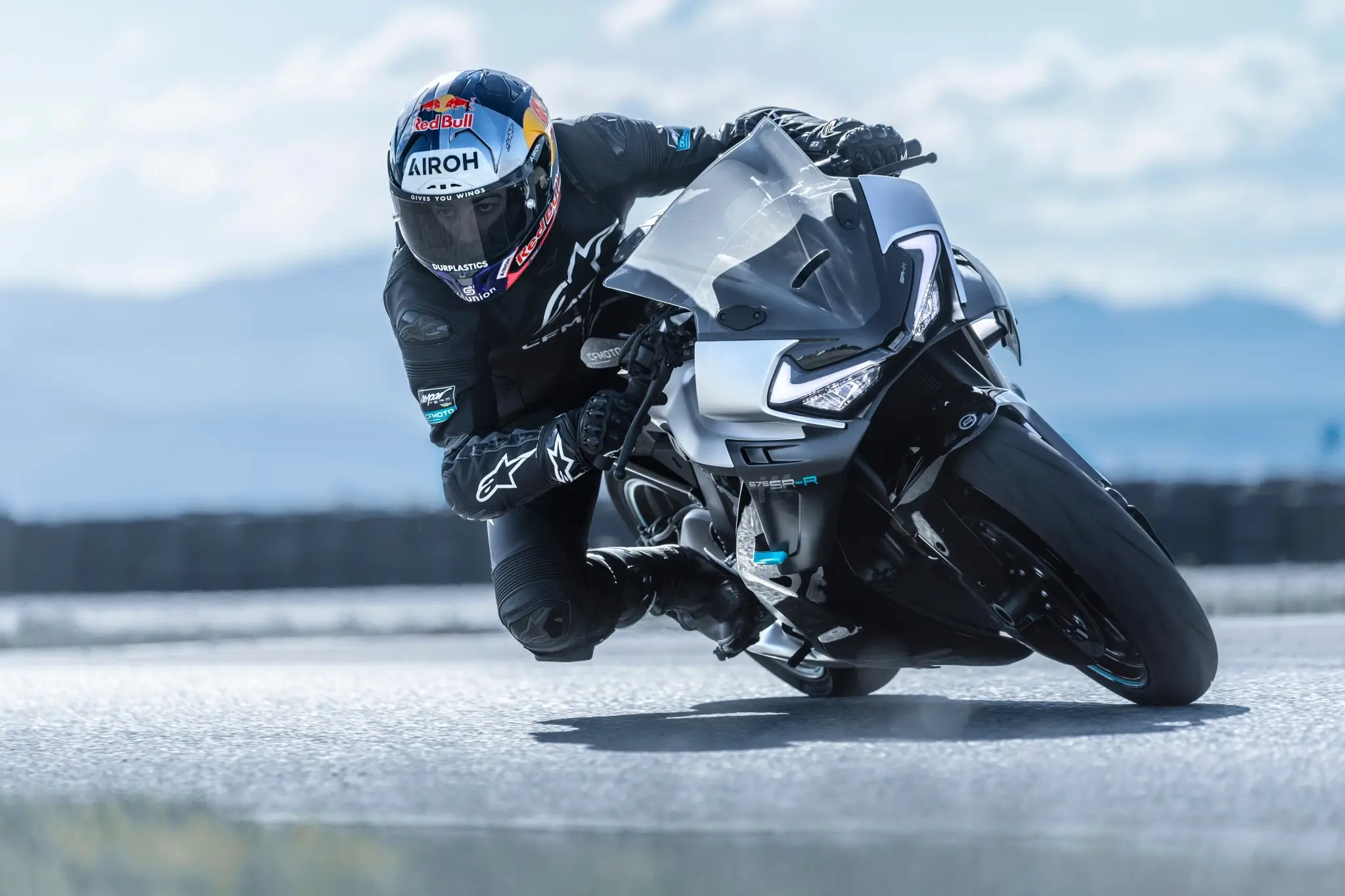 A motorcyclist in full racing gear leans sharply into a turn on a 675SR-R sport motorcycle on a paved track.