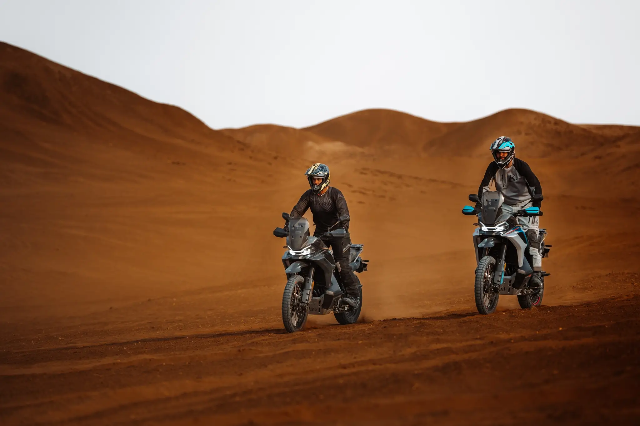 Two people wearing helmets and gear ride 800MT-X motorcycles across a sandy desert landscape, with rolling dunes in the background.