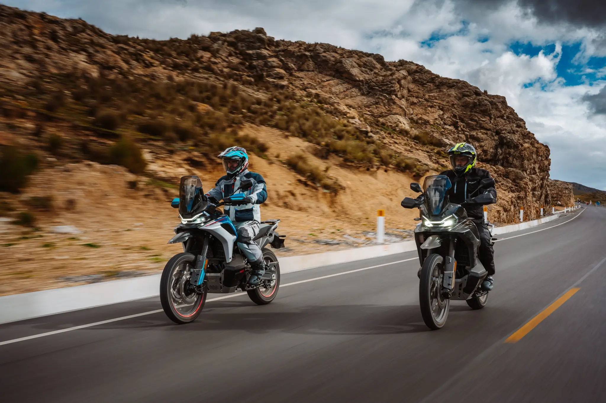 Two people wearing helmets and riding gear ride 800MT-X motorcycles on a paved road through a rocky, mountainous landscape under a cloudy sky.
