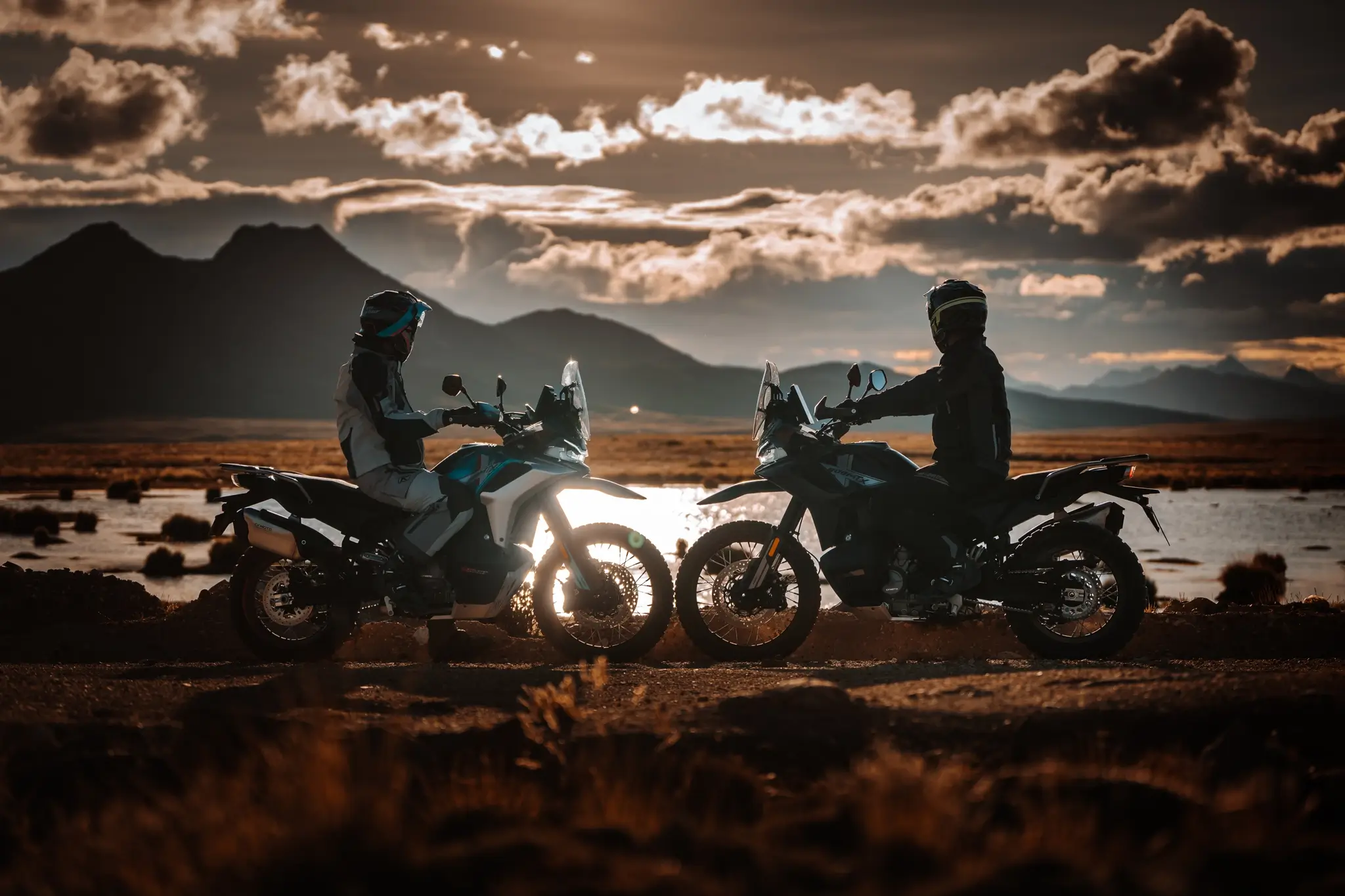 Two people wearing helmets sit on 800MT-X adventure motorcycles facing each other on a dirt road, with mountains and a cloudy sunset sky in the background.