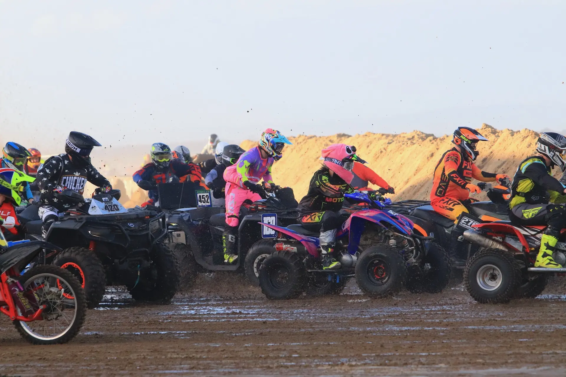 A group of ATV riders wearing colorful racing gear compete on a muddy dirt track, with dirt and debris flying in the air—evoking the energy of the Weston Beach Race and showcasing top brands like CFMOTO.