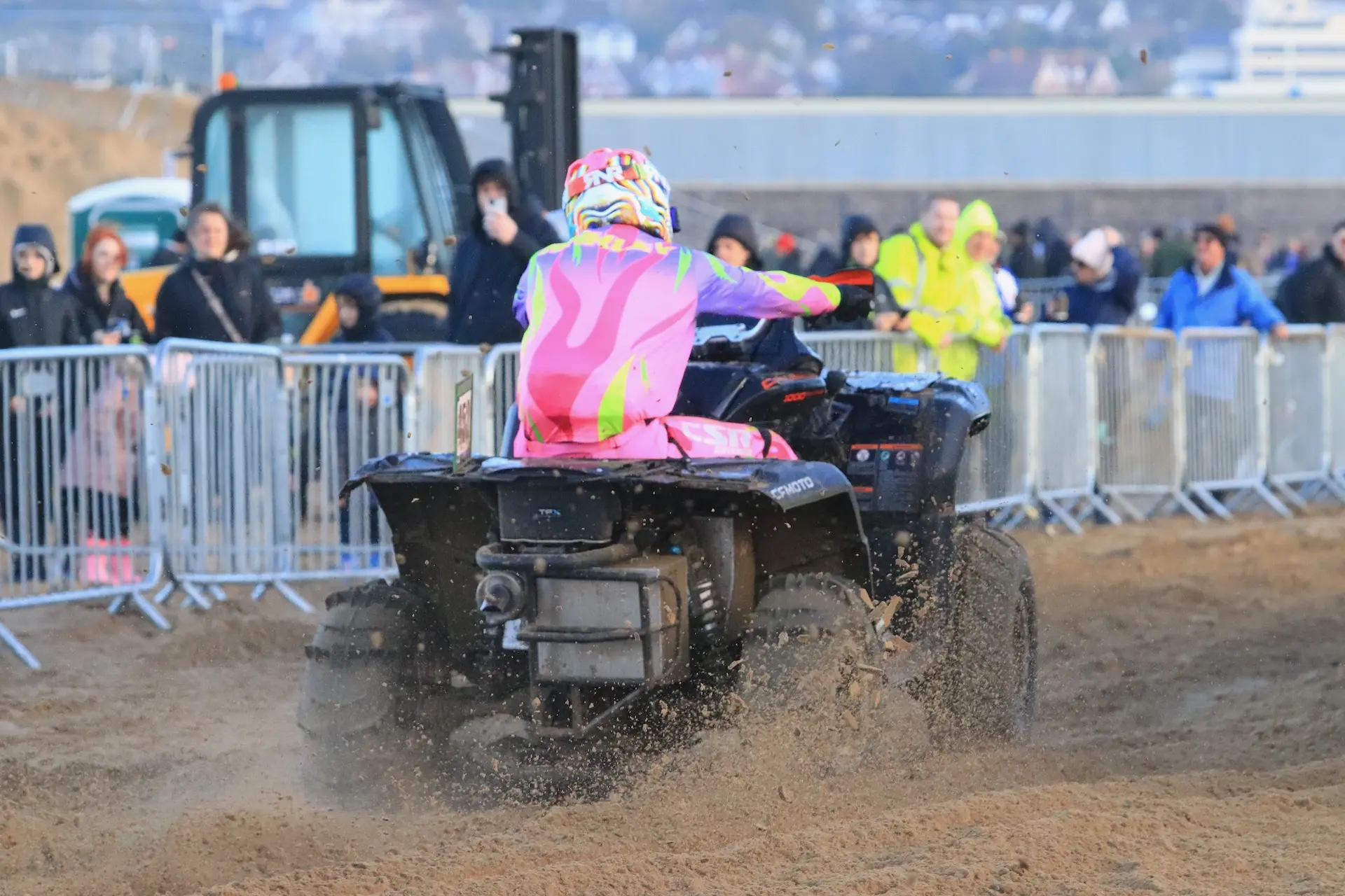 Sporting a colorful outfit, Harry Hinkley rides a CFMOTO ATV on the sandy track at the Weston Beach Race, with spectators and barricades in the background.
