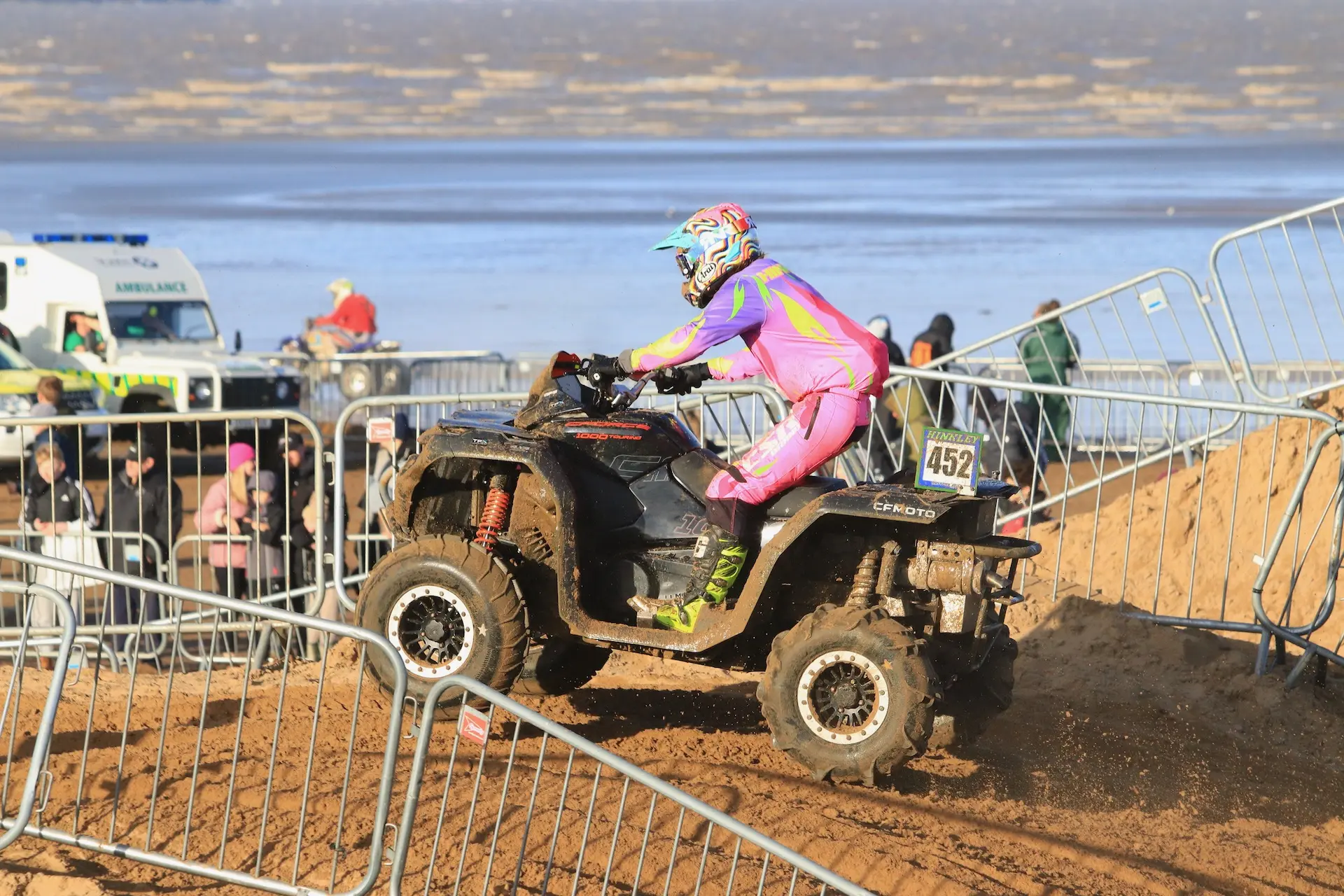 A person in a colorful suit rides a CFMOTO ATV up a sandy incline during the Weston Beach Race, with spectators and an ambulance visible in the background.