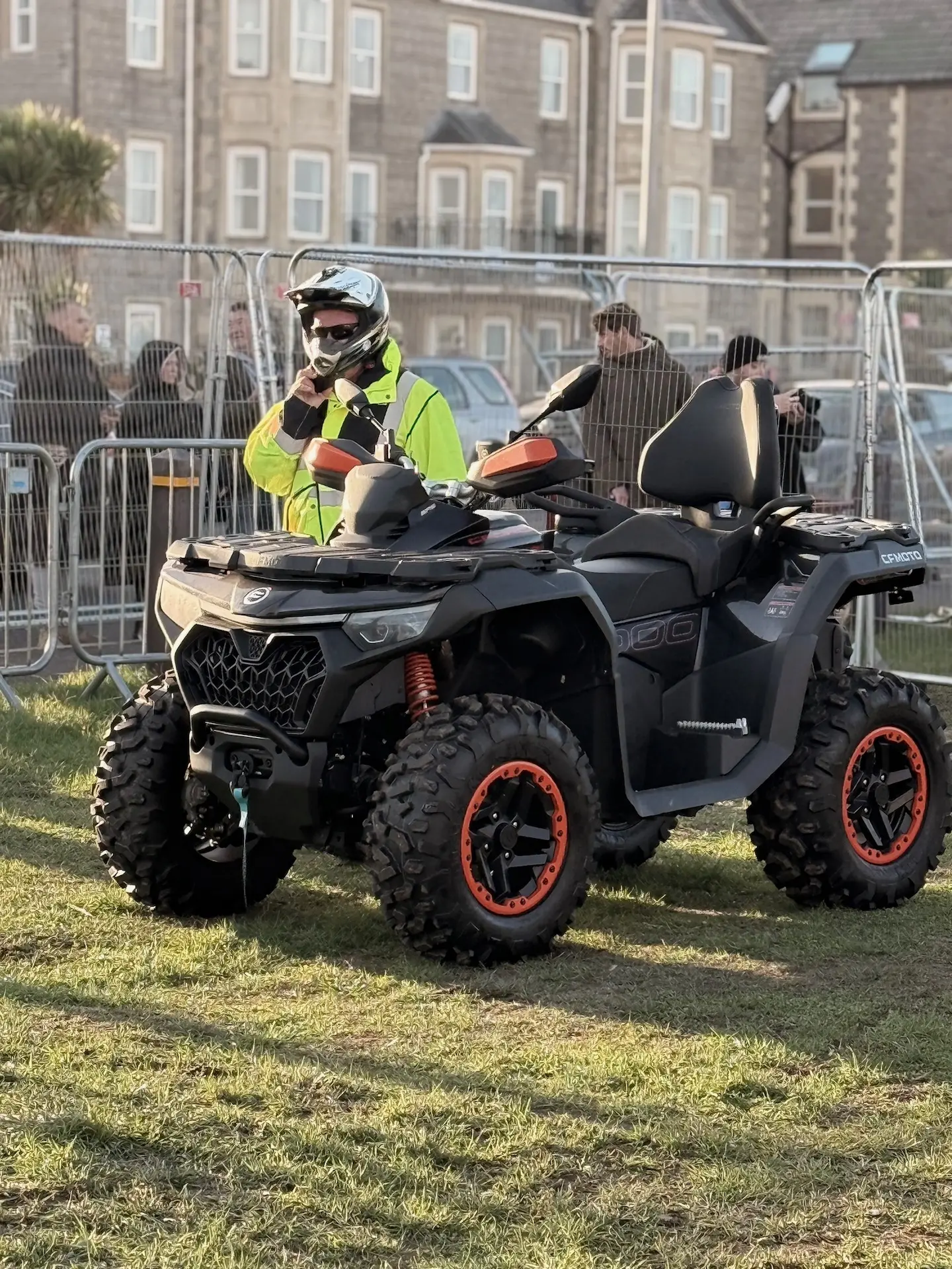 A person in a reflective jacket and helmet stands beside a black and orange CFMOTO all-terrain vehicle on grass, with metal barriers and buildings in the background, possibly at the Weston Beach Race.