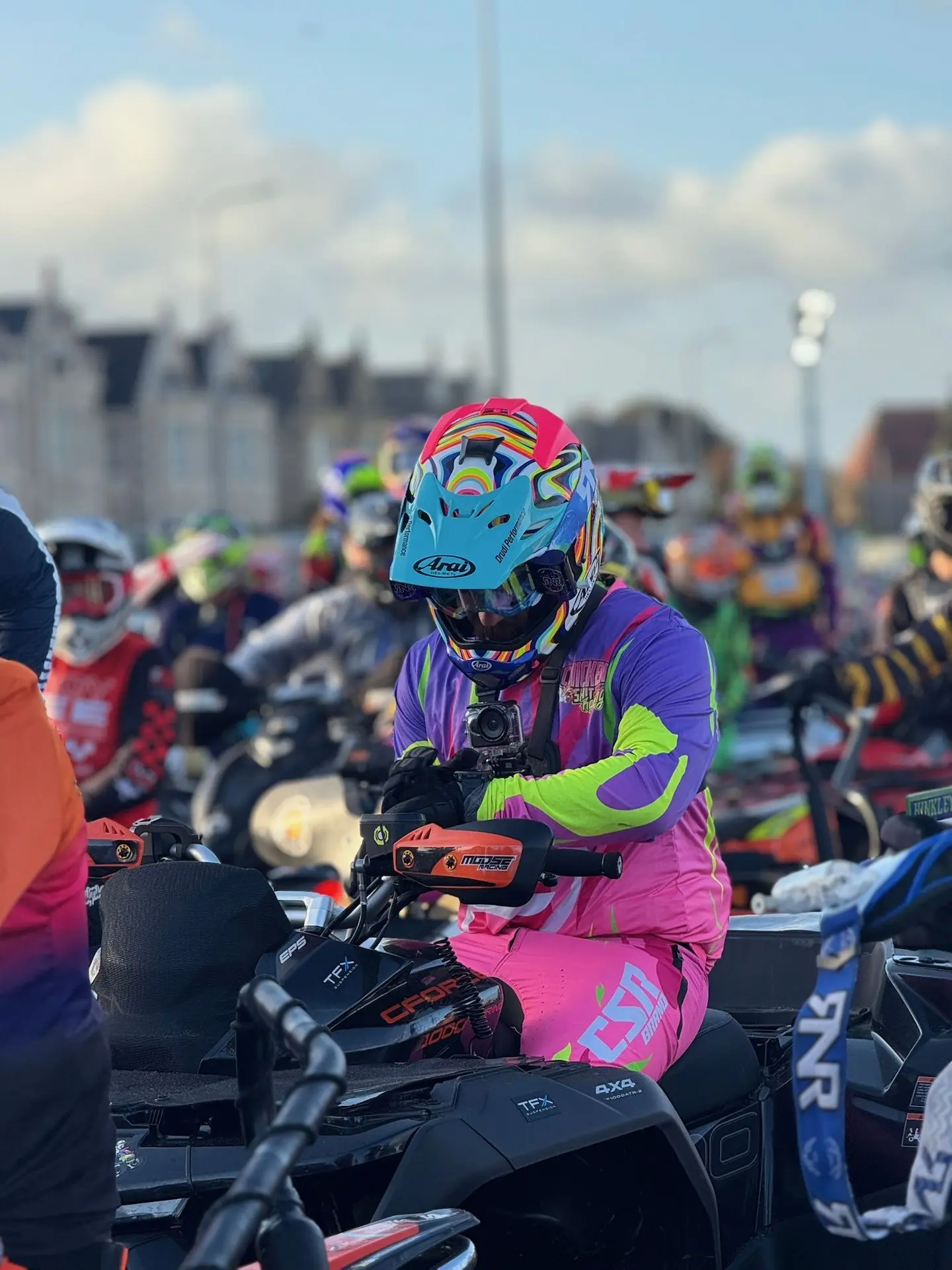 A group of motocross riders in colorful gear and helmets prepare for a race, with one rider in pink and purple gear sitting on a CFMOTO ATV in the foreground, ready for the Weston Beach Race.