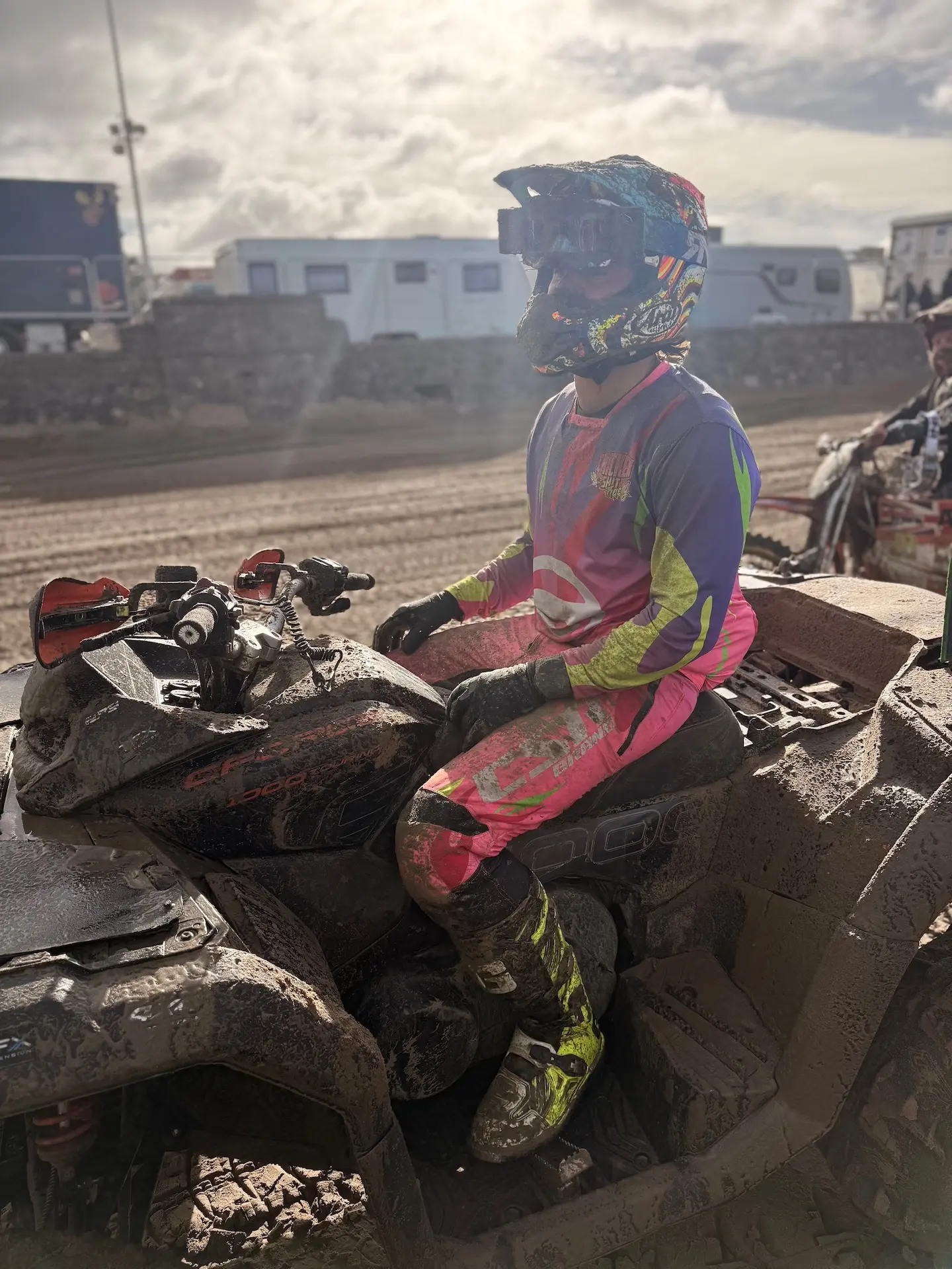 A person in colorful motocross gear and helmet sits on a muddy CFMOTO ATV at an outdoor track, with vehicles and RVs visible in the background—capturing the thrill of events like the Weston Beach Race.