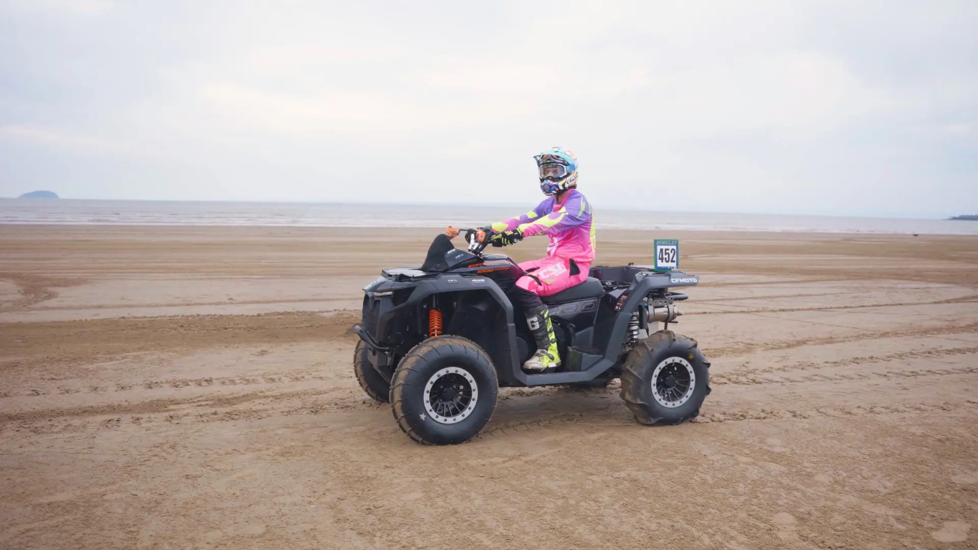A person wearing a colorful racing suit and helmet rides a CFMOTO ATV on a sandy beach with overcast skies, reminiscent of the thrilling Weston Beach Race.
