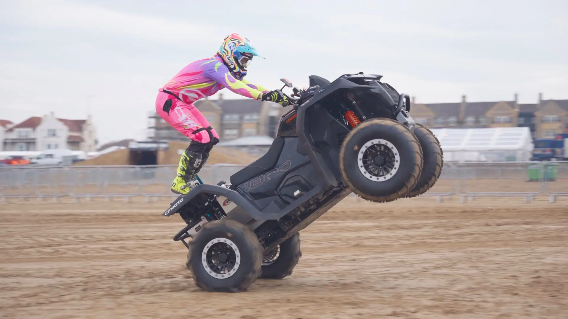 A person in colorful gear performs a wheelie on a CFMOTO all-terrain vehicle (ATV) on a sandy surface at the Weston Beach Race, with buildings in the background.
