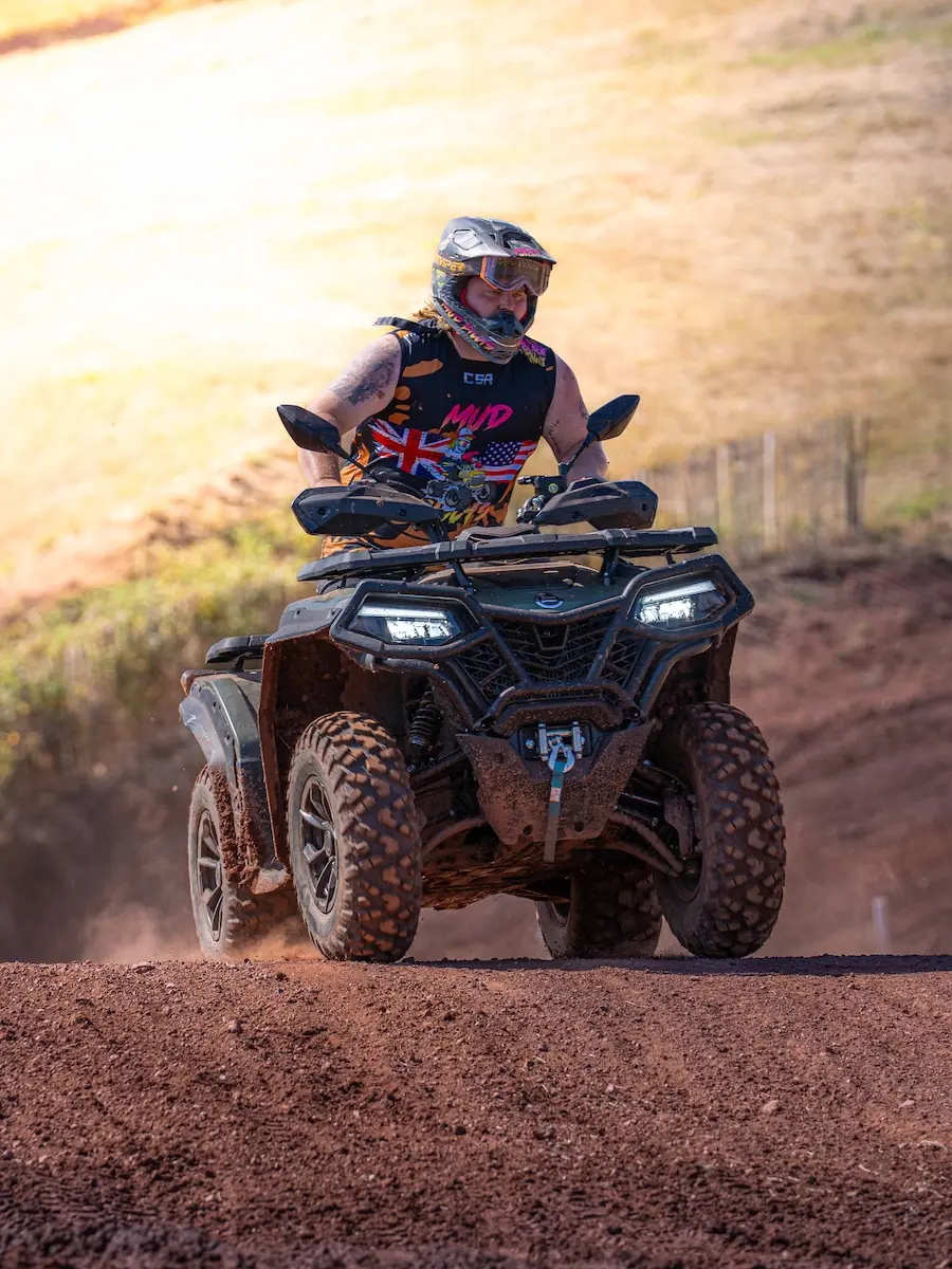 A person wearing a helmet and protective gear rides a CFMOTO CFORCE 1000 ATV on a dirt track, with dust and dirt coating the vehicle's tires in the 4x4 class.