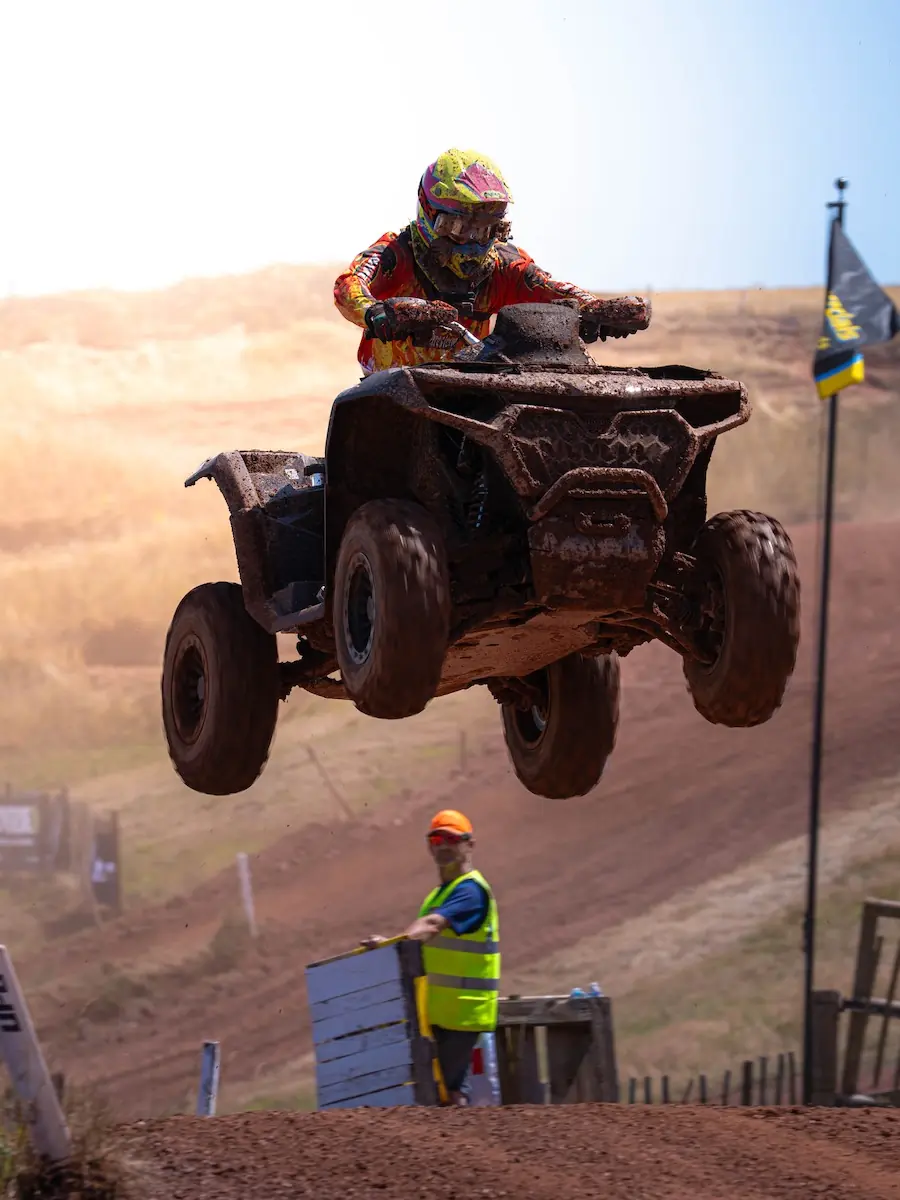 A person riding a CFMOTO CFORCE 1000 ATV soars airborne over a dirt track during a 4x4 class race, with a safety official and flag visible in the background.
