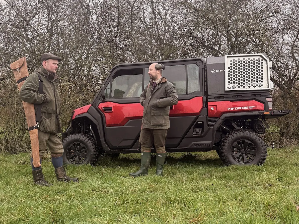 Two men in outdoor attire stand beside a red UFORCE U10 PRO XL off-road utility vehicle with a dog crate attachment, on a grassy area near leafless bushes.