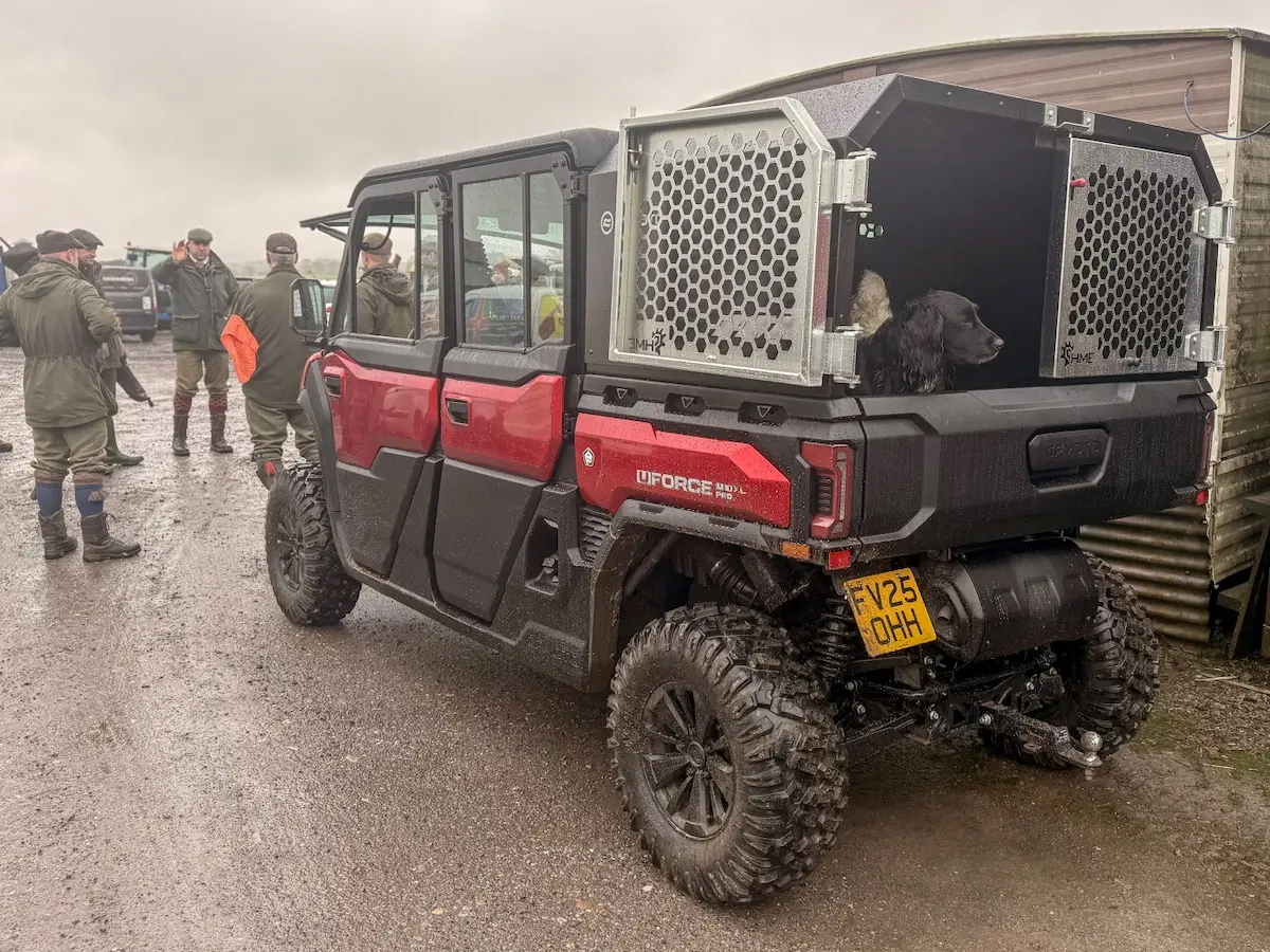 A red UFORCE U10 PRO XL off-road utility vehicle with a dog in a rear cage is parked on a muddy road near several people standing and talking.