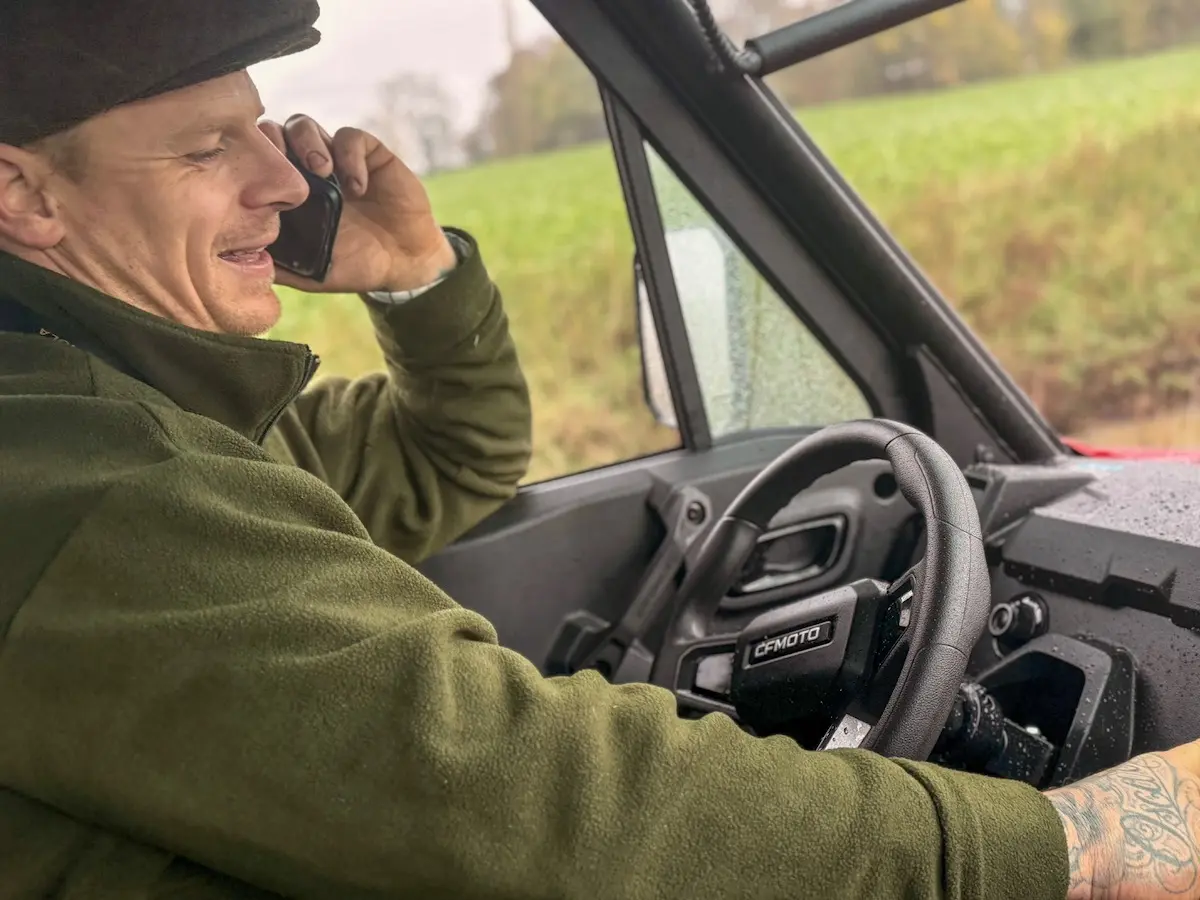 Man in a green jacket and cap talking on a phone while sitting in the driver's seat of a UFORCE U10 PRO XL, with a tattoo visible on his right wrist.