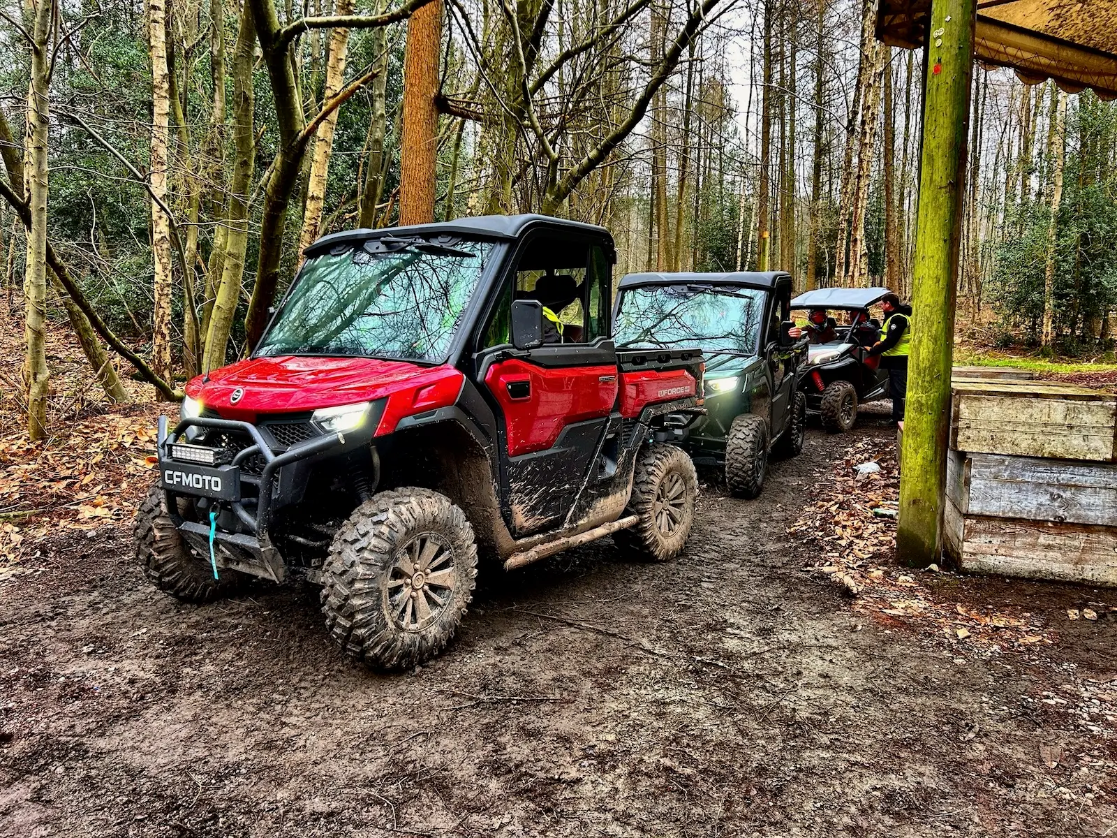 Three farm quads are parked in a muddy forest area, partially sheltered by a wooden structure.