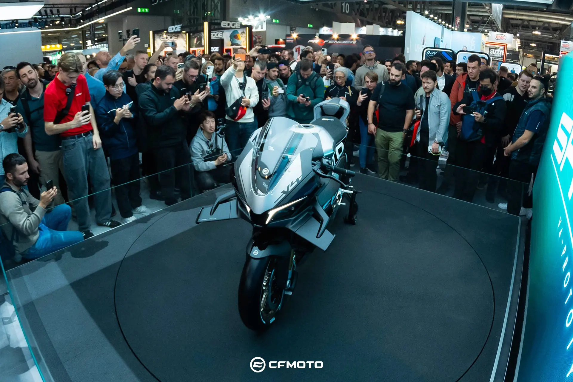 A crowd observes and photographs the CFMOTO CFORCE TECH GEN 4 Concept sport motorcycle displayed on a circular platform at an indoor event.