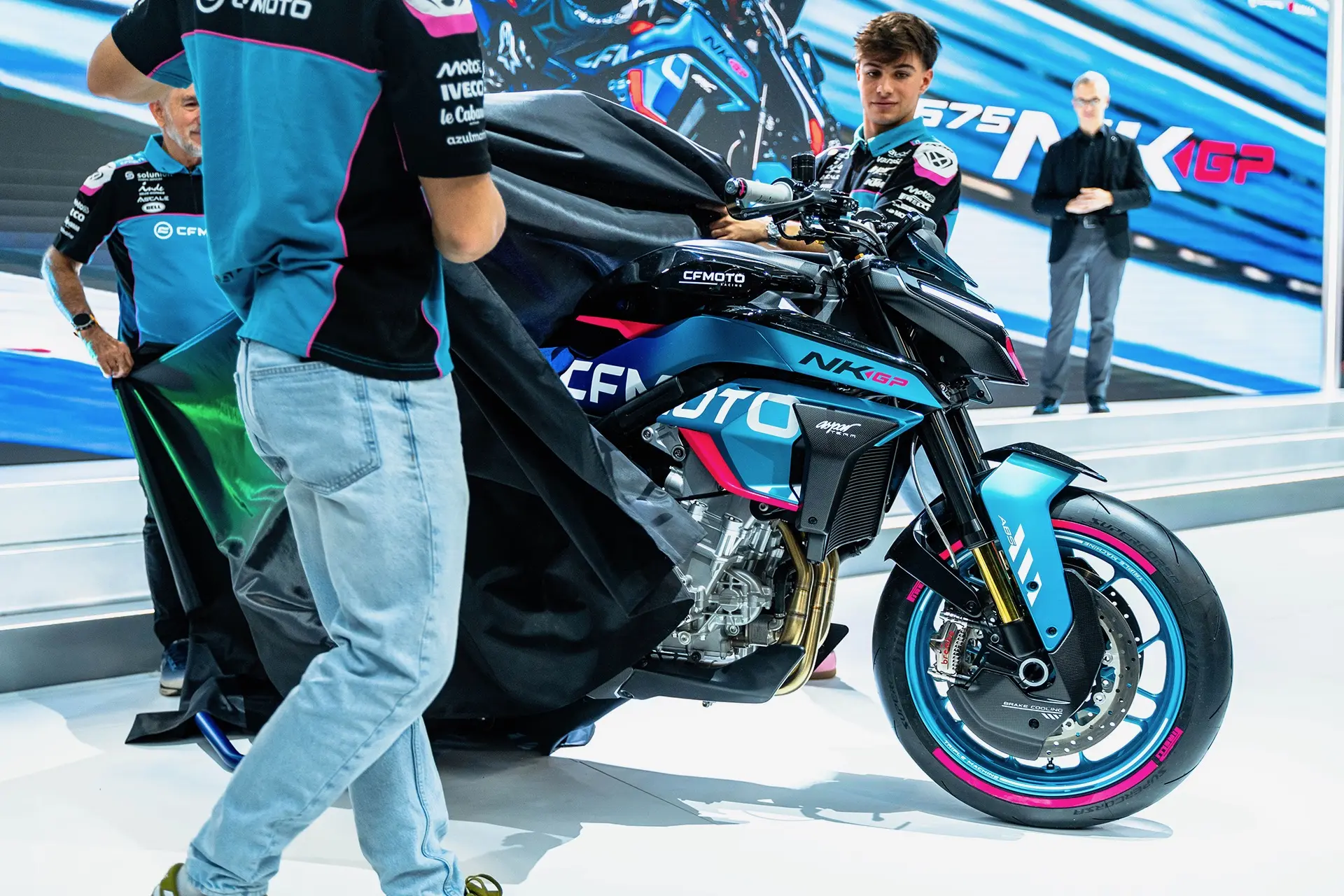 Team members unveil the CFMOTO V4 SR-RR Prototype motorcycle at an event, with striking backdrop graphics and a person observing in the background.