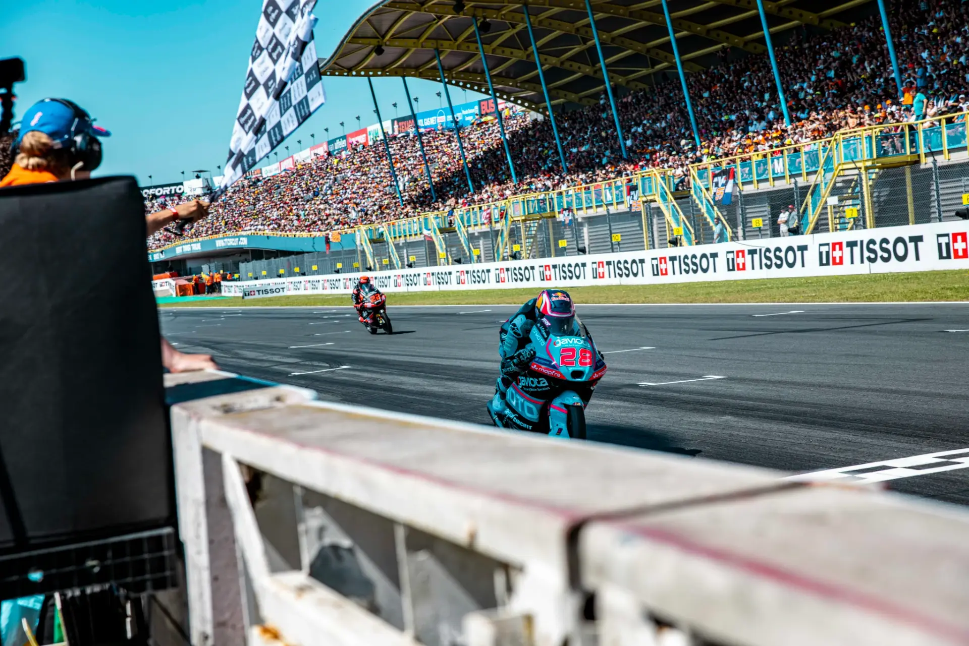 A motorcycle racer crosses the finish line at the Dutch MotoGP on a CFMOTO bike as a chequered flag waves, with Quiles and Alonso competing fiercely, a large crowd in the stands, and another rider in the background.