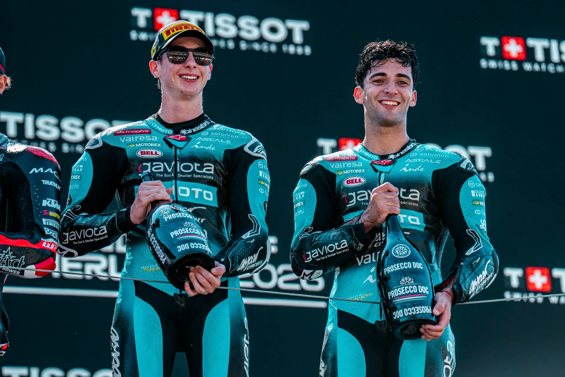 Two motorcycle racers in teal and black CFMOTO suits stand on a podium, each holding a large bottle of prosecco, with a Tissot logo backdrop behind them at the Dutch MotoGP Grand Prix.