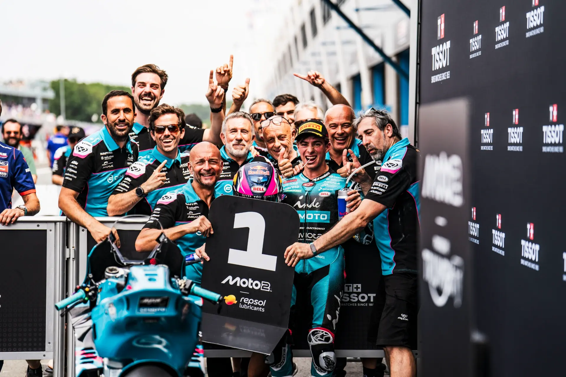 A motorcycle racing team celebrates with a CFMOTO rider holding a "1" sign, indicating first place, in front of a Grand Prix racing circuit backdrop.