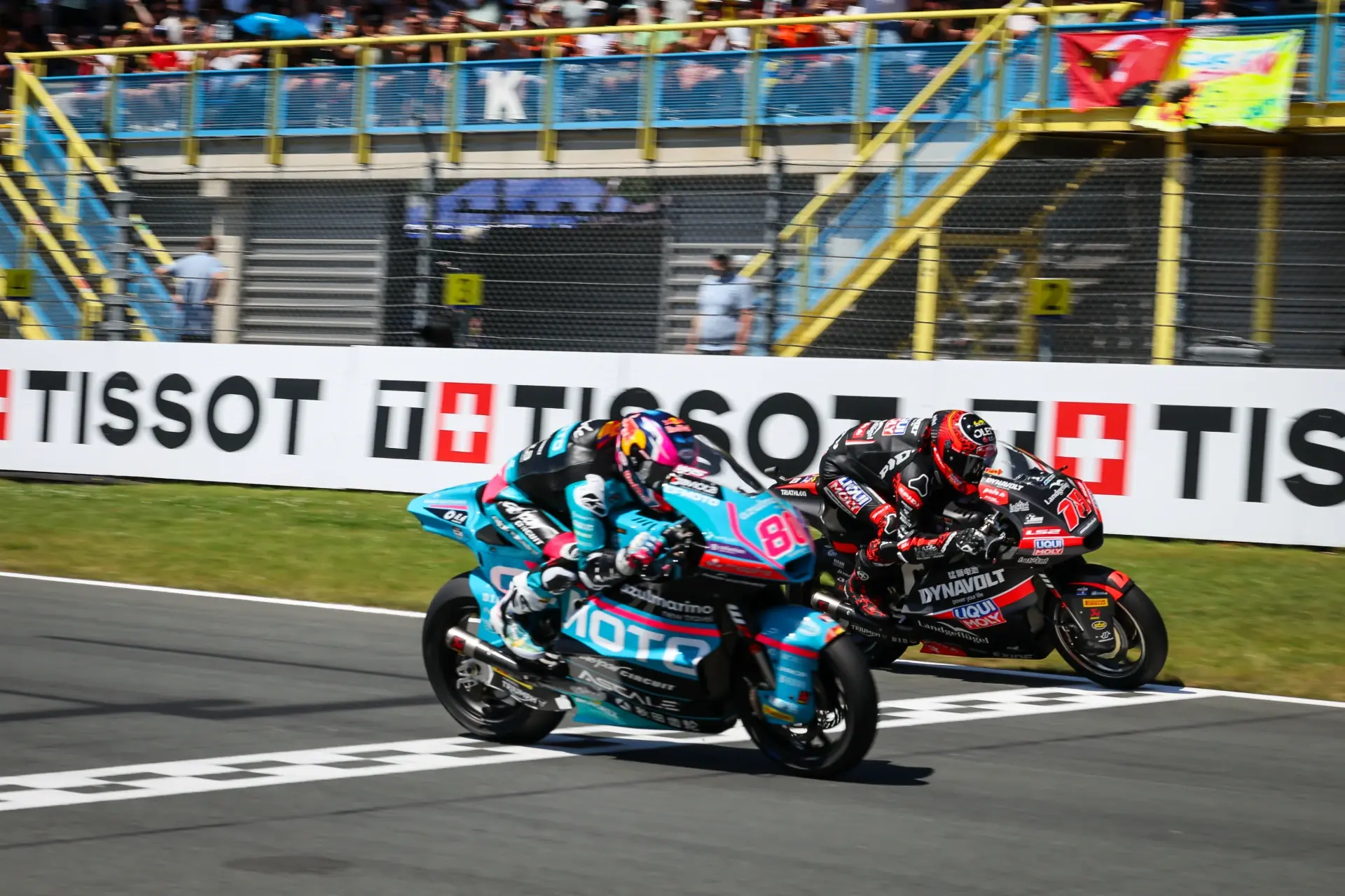 Two motorcycle racers cross the finish line side by side on a Grand Prix racetrack, with spectators watching from the stands in the background.