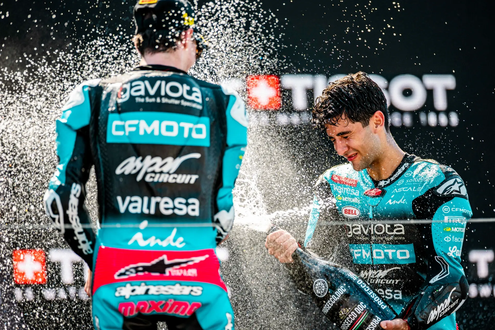 Two motorcycle racers in teal racing suits celebrate on the podium, spraying champagne after the Dutch MotoGP Grand Prix, with CFMOTO sponsor logos in the background.