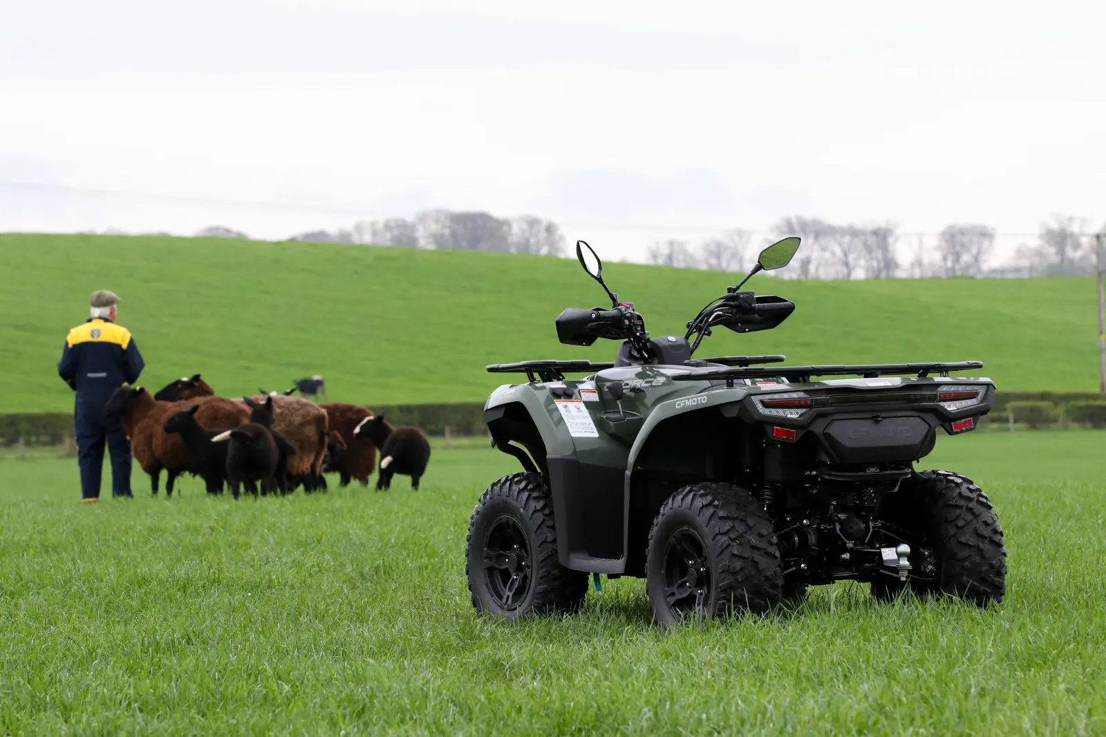 A CFMOTO CFORCE TECH GEN 4 Concept quad bike is parked on grass in the foreground while a person stands near a group of cows in a green field in the background.