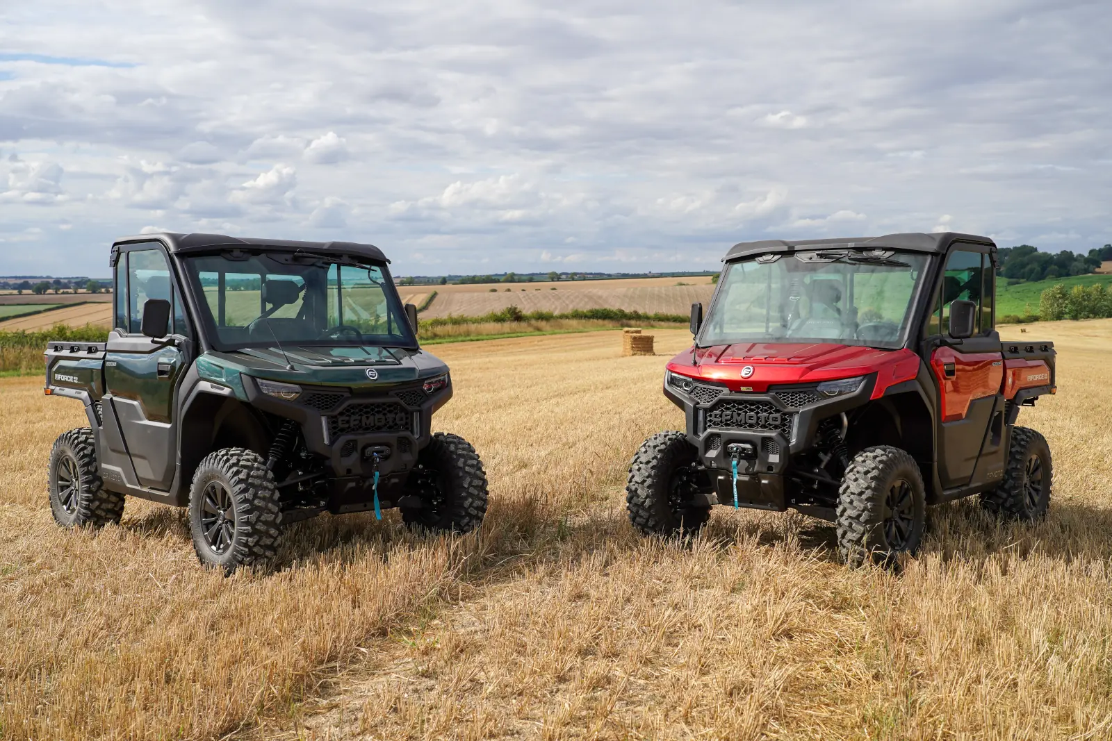 Two off-road utility vehicles, including a black CFMOTO CFORCE TECH GEN 4 Concept and a red model, are parked side by side in a harvested field under a cloudy sky.