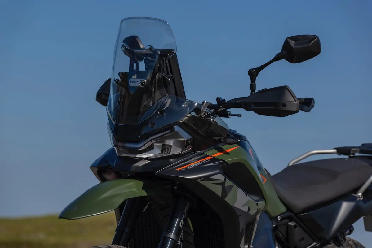 Close-up of a black and green adventure motorbike, featured in our CFMOTO 1000MT-X review, with a tall windscreen, side mirrors, and angular bodywork set against a clear blue sky.