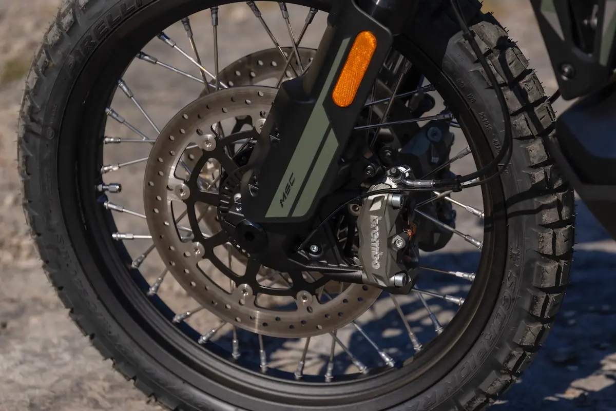 Close-up of a CFMOTO 1000MT-X front wheel showing a disc brake, black spokes, rugged tyre, and Brembo brake calliper on a sandy surface—perfect for any detailed CFMOTO 1000MT-X review.