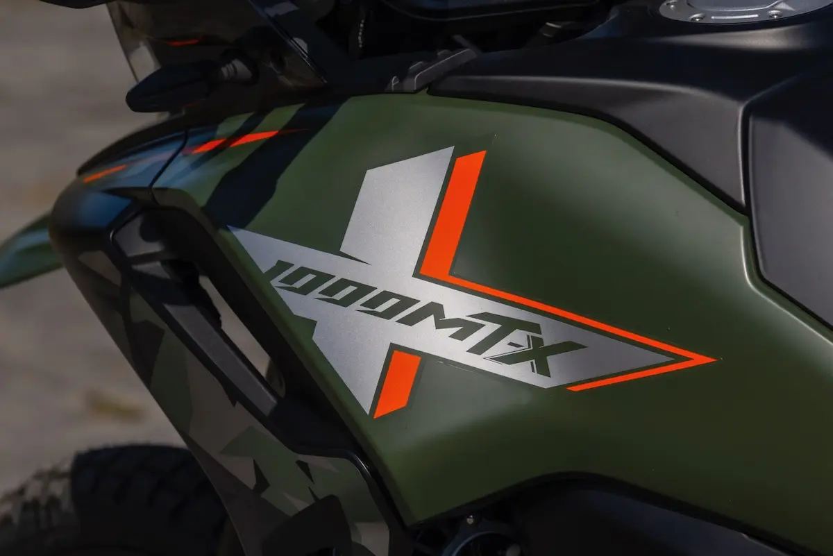 Close-up of a motorcycle tank with a matte green finish, featuring "1000MT X" graphics in silver, orange, and black—perfect for your next CFMOTO 1000MT-X review.