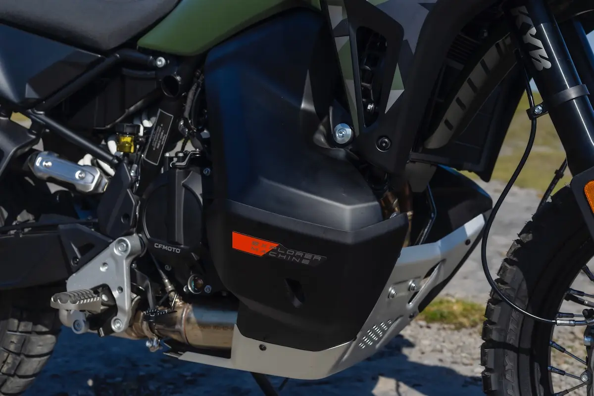 Close-up of a motorcycle engine and side panel with a "CFMOTO" label and visible protective sump guard, photographed outdoors—perfect for a CFMOTO 1000MT-X review.