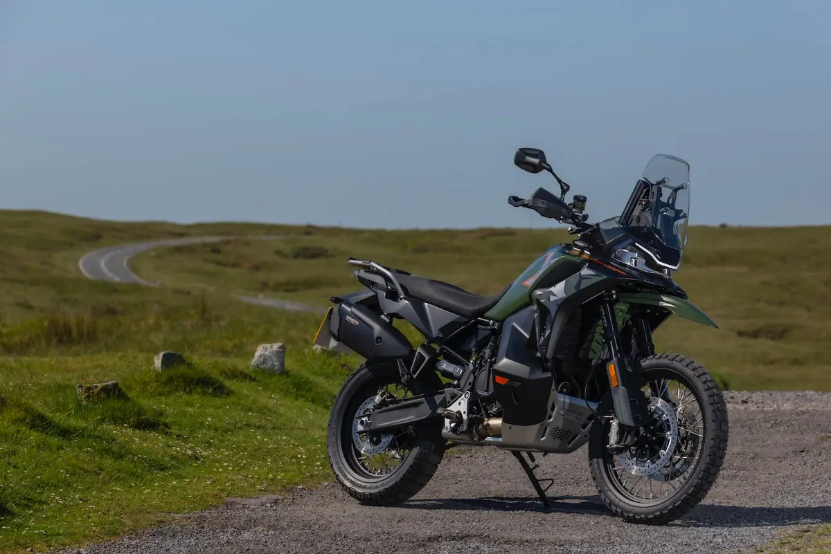 A green and black adventure motorbike, featured in a recent CFMOTO 1000MT-X review, is parked on gravel beside an open grassy landscape with a winding road in the background under a clear sky.