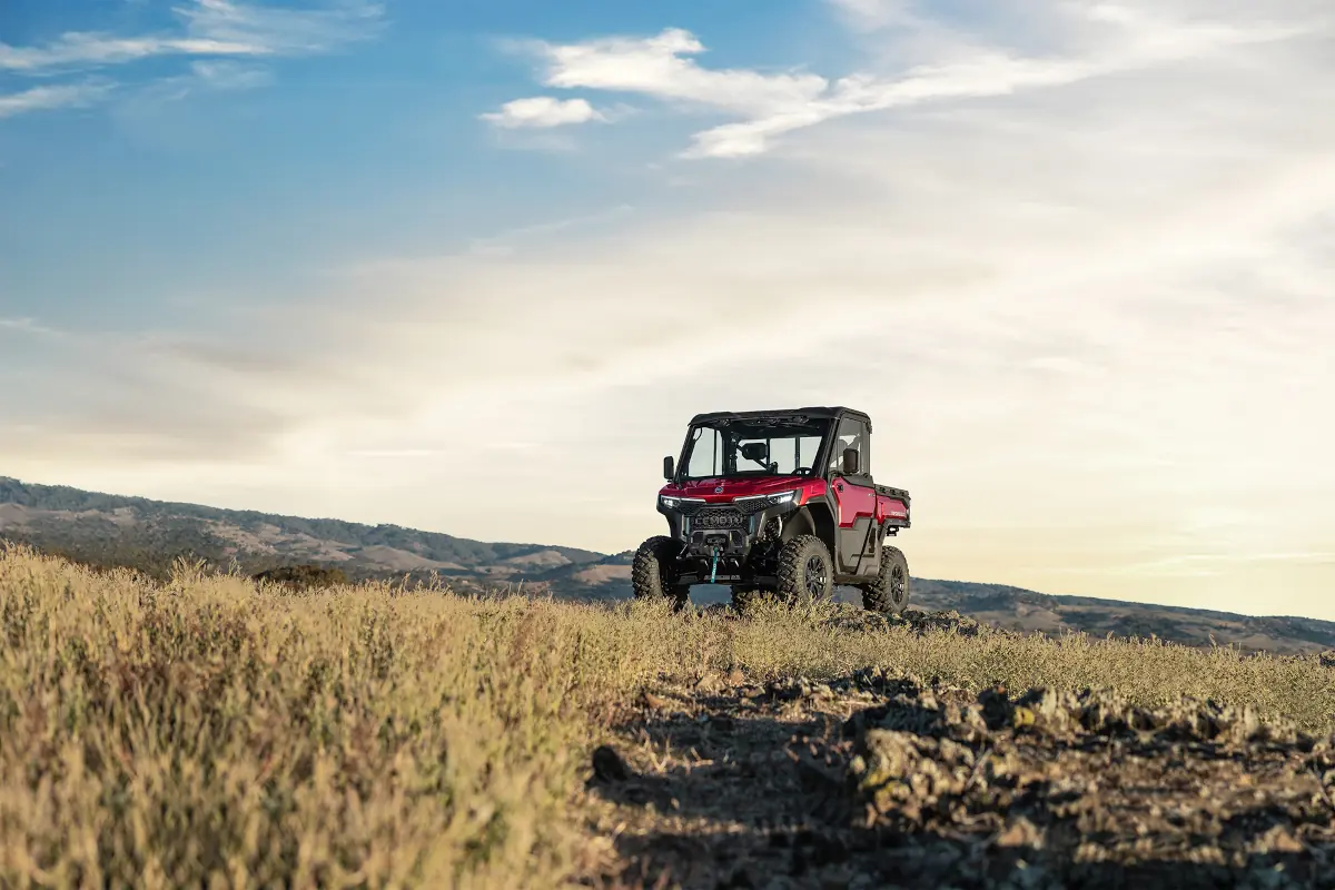 A red off-road utility vehicle, commonly used in agriculture, is parked on grassy terrain with hills and a partly cloudy sky in the background.