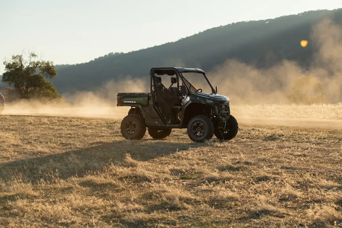 A black UFORCE U10 PRO AGRI utility terrain vehicle drives across a dry, grassy field with dust trailing behind, set against a backdrop of hills and sunlight.