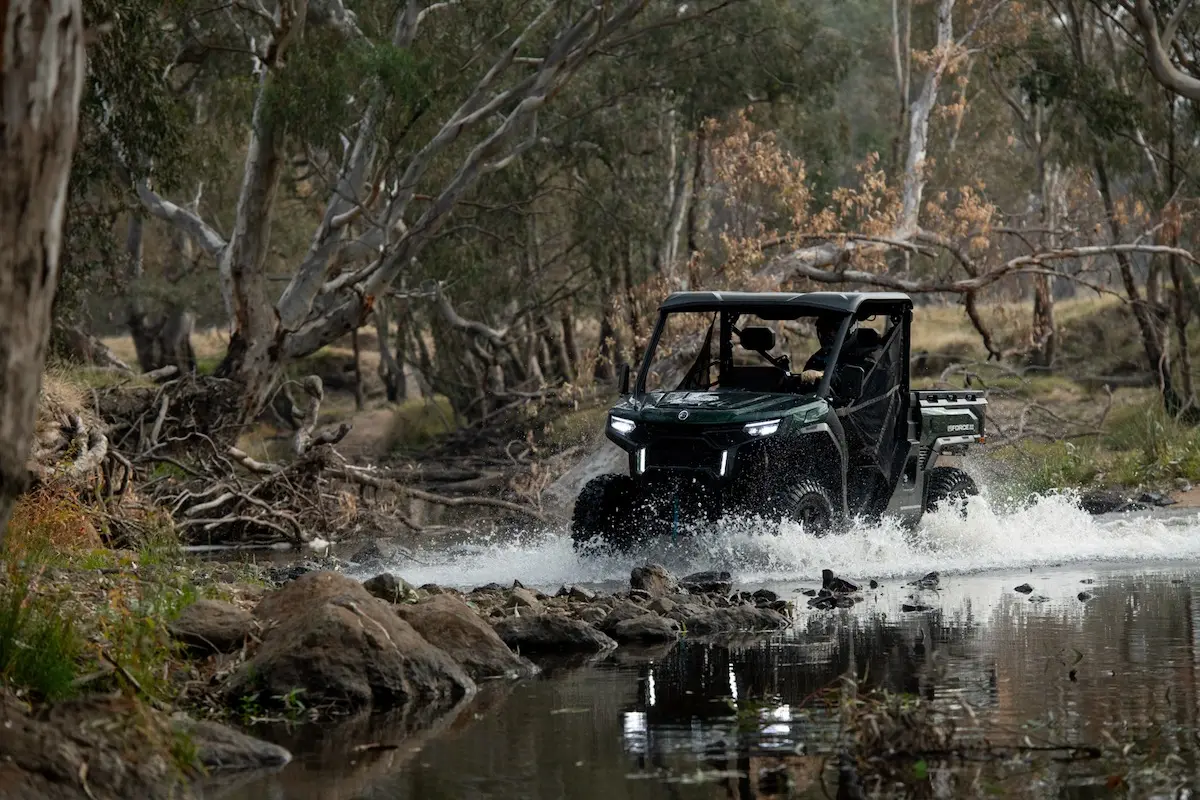 A UFORCE U10 PRO AGRI off-road utility vehicle drives through a shallow, rocky creek surrounded by trees and natural vegetation.