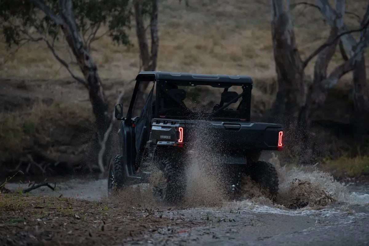 A black UFORCE U10 PRO AGRI off-road utility vehicle drives through a shallow, muddy creek, splashing water, with trees and dry grass in the background.