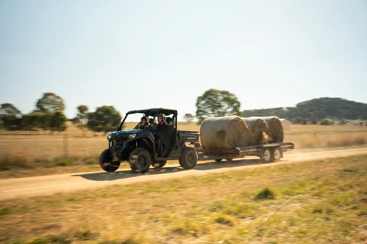 A UFORCE U10 PRO AGRI utility vehicle tows a trailer loaded with large hay bales down a dirt road in a rural area.